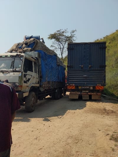 The scene depicts a daylight view of an unpaved, sandy dirt road in Rutshuru, Democratic Republic of the Congo. Two heavy-duty cargo trucks are stationary on the road.
In the lower left foreground, the back of a person wearing a dark red shirt is partially visible, facing away from the camera.
The truck on the left has a white cab and a multi-axle chassis, carrying a substantial, uneven load covered predominantly by blue and brown tarpaulins. A blue barrel is secured on top of the cargo near the cab. This truck is angled slightly towards the right, appearing to be positioned behind the second truck.
To its immediate right, the rear of a dark blue box-body truck is visible. This truck displays partial markings on its rear, including "L.P.D." and "T.R.C.", along with other indecipherable script. The trucks appear to be positioned in close proximity, suggesting a stop or a narrow passage.
The road surface is unpaved, composed of light brown sand and dirt, with visible tire tracks and scattered small stones. In the background, a steep, sparsely vegetated hillside rises on the right. A lone, small tree with bare branches stands between the two vehicles, silhouetted against a clear, light blue sky.