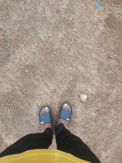 An overhead, full-frame shot shows a person's feet and lower legs standing on a rough, sandy surface in Ajasse Ipo, Nigeria. The person is wearing dark gray slide sandals with a textured strap and black pants. A yellow fabric is visible at the bottom of the frame, obscuring the lower portion of the person's body. The ground is composed of small pebbles and sand, with a few scattered small rocks. A single, light-colored rock, roughly triangular in shape, rests on the ground to the right of the person's feet. There are a few dried grass blades near the right sandal. The overall environment appears to be an outdoor, possibly unpaved area.