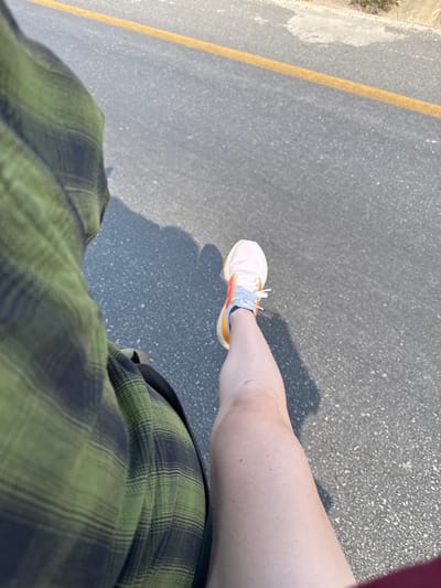 A top-down shot captures a person's lower body on asphalt. The person wears a green and black plaid shirt, and a running shoe with white, orange, and yellow accents is visible on their right foot. A shadow is cast on the asphalt near the shoe, and a yellow line runs along the edge of the pavement. The person is likely walking on a road or sidewalk.