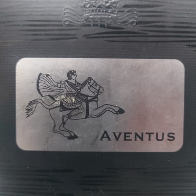The image depicts a close-up, top-down view of a dark-colored object, likely a container or packaging, featuring a prominent metallic label. The object's main body is dark, possibly black, with a textured surface characterized by horizontal raised ridges. At the very top, a partially visible, dark, embossed emblem or crest featuring wavy lines and a central motif is present.

Centered on this dark background is a rectangular, silver-toned metallic label with rounded corners. The label displays a black outline drawing of a man riding a rearing horse, facing towards the right. The rider wears what appears to be classical attire, including a flowing cape and a pleated tunic or skirt. Below and to the right of the horse figure, the word "AVENTUS" is printed in black, uppercase sans-serif letters. The metallic surface of the label exhibits numerous superficial scratches and scuff marks, particularly around the horse illustration, indicating wear.

The image is a static close-up of an object and does not contain visual information to determine a specific location within Gimpo-si, South Korea, or any other urban environment. No people are visible beyond the depicted illustration. No specific actions or interactions are depicted apart from the static representation of a man on horseback.