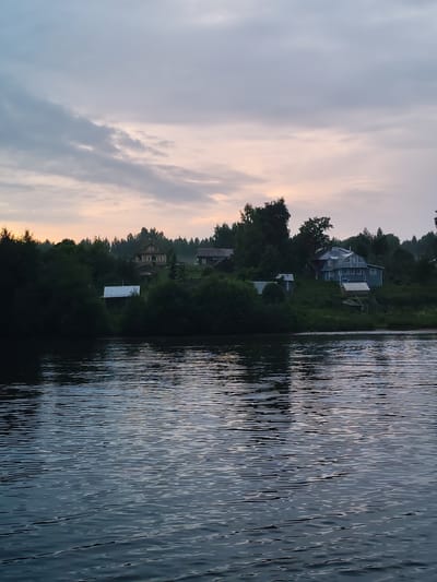 The image depicts a tranquil waterscape with houses nestled on a tree-lined hill overlooking the water. The sky is overcast with hues of gray and soft pink, suggesting either dawn or dusk. The water is dark and rippled, reflecting the subdued light of the sky and the silhouettes of trees and buildings. Several houses are visible on the elevated bank, with varying architectural styles, including a prominent light blue house with multiple windows and a gabled roof, and a more traditional yellow wooden house. The surrounding vegetation is dense and green, with tall trees dominating the landscape. The scene appears to be a quiet rural or semi-rural setting. The location is described as Ratibor, Russia. There are no visible people or animals in the image, and no discernible actions or interactions are occurring.