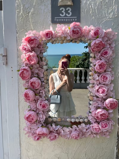 A woman is reflected in a mirror decorated with pink flowers and seashells. The mirror is affixed to a textured wall. Above the mirror is a sign with the number "33" and Cyrillic text. In the reflection, the woman holds a pink phone, taking a selfie. She wears a white dress and carries a light blue handbag. The background in the reflection shows a body of water, likely the Black Sea, with a white railing and green foliage. The scene is located on Primorska Street in Kryzhanivka, Ukraine.
