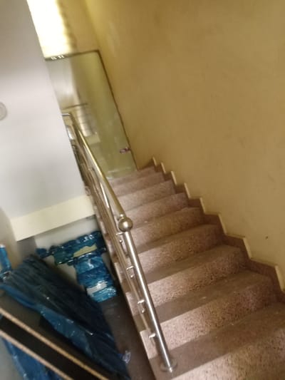 This media file depicts an indoor staircase with a stainless steel railing. The staircase treads appear to be made of speckled concrete. To the left of the stairs, there are several large items wrapped in blue plastic sheeting, possibly construction materials or furniture. The walls are painted a light beige color.
The scene is brightly lit, suggesting it is daytime. The overall impression is of a functional, possibly residential or commercial building's interior. There are no people visible in the frame, and no text is apparent. The image appears to be taken at an upward angle, looking up the staircase. The building's interior design is simple and utilitarian. The presence of the plastic-wrapped items suggests ongoing construction, renovation, or storage.