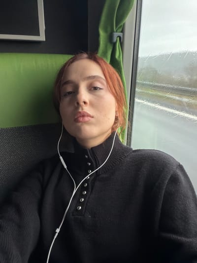 The scene depicts a young adult female seated inside a moving vehicle in Moineville, France. She has reddish-brown hair and a light complexion, looking directly at the camera with a neutral expression. She is wearing a black, long-sleeved knitted sweater featuring a high collar secured by five visible silver snap buttons down the front placket. White wired earphones are inserted into both ears, with the cable extending down her chest and an inline remote control unit visible. To her left, part of a green fabric seat or headrest is visible, along with a dark rectangular screen or panel. To her right, a green fabric curtain is pulled back by a grey plastic clip, revealing a rain-streaked window. Through the window, a blurred, overcast, and wet exterior environment, possibly a highway with faint white lane markings, is visible, indicating motion and precipitation.