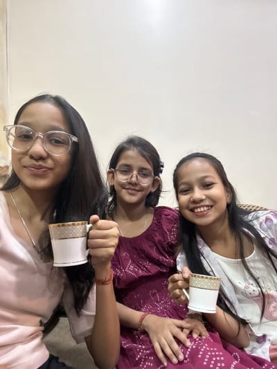 Three young women are angled in the frame, each holding a white mug with a gold design. The top girl wears a light-colored patterned top and smiles widely. The middle girl wears glasses and a dark patterned top, also smiling. The bottom girl, closest to the camera, sports clear-framed glasses and a light pink top. They appear to be indoors.