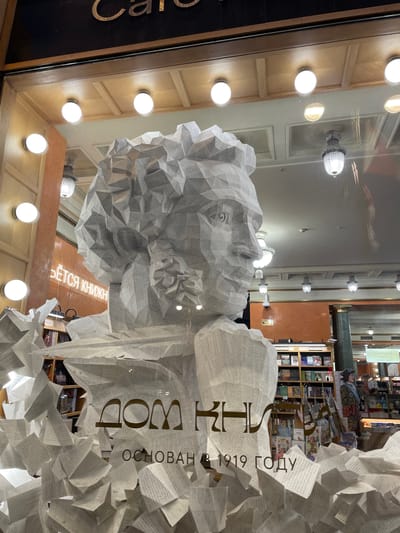 The image shows a bookstore window display featuring a large sculpture made of paper, resembling a bust of a person with intricate details. The sculpture is surrounded by scattered paper elements, giving a sense of disarray and artistic expression. Behind the sculpture, shelves filled with books are visible, indicating the setting is a bookstore. Text on the window reads "ДОМ КНИГИ" (House of Books) and "ОСНОВАН В 1919 ГОДУ" (Founded in 1919). The overall environment appears to be indoors, likely a commercial establishment. The location of the city of Kovalevo, Russia is not discernible from the image itself.
