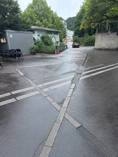 This image captures an outdoor scene, likely a street or service road, in Liestal, Switzerland, under an overcast sky following rain, as indicated by the wet asphalt and damp surfaces. The ground features dark, wet asphalt with distinct white road markings, including several sets of triple dashed lines and a single solid line. Embedded concrete blocks form linear patterns and an "X" intersection across the asphalt, with some areas showing cracks and repairs.
On the left side of the frame, a dense, green hillside covered with trees and foliage forms a lush backdrop. In the mid-ground, a dark-colored car, possibly an SUV or hatchback, is visible with its illuminated tail lights, partially obscured by the trees. Further in the background, several multi-story buildings with lights visible through windows suggest an urban or suburban setting.
Closer to the foreground on the left, there is a light-colored building with a green roof, surrounded by green bushes. A blue sign featuring a white wheelchair symbol (handicap accessibility) is affixed to its wall. Nearby, two orange and white traffic cones stand on the wet ground. Adjacent to this, a dark grey, modern-style building, possibly a kiosk or small commercial unit, has several black chairs stacked neatly outside. A small screen on this building displays colorful, indistinct graphics, and faint text, possibly "CURVO," is visible on its side. A concrete wall forms a barrier on the far upper left. The overall impression is a quiet, overcast daytime scene.