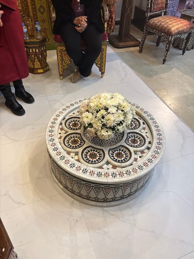 A round table with intricate mosaic patterns sits on a white marble floor, topped with a large bouquet of white chrysanthemums. Around the table are ornate, carved wooden chairs. Two people are partially visible, one in black pants and beige shoes, and another in a red coat and black boots. The scene appears to be in a formal interior setting, possibly a museum or cultural center.