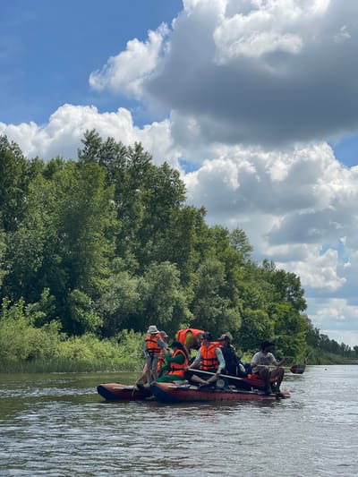 On a cloudy day, a group of people are on two rafts in a wide river, with a dense green forest on the far bank. The rafts are red and orange, and most of the people are wearing orange life vests. Some individuals are holding paddles, suggesting they are engaged in a recreational activity like rafting or kayaking. The water appears calm, with gentle ripples. The sky is a mix of blue sky and large, fluffy white and grey clouds. The vegetation on the bank is lush and varied, with tall trees and smaller shrubs. The scene suggests a summer day spent outdoors in a natural setting.