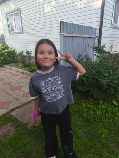 A young girl with long dark hair is posing for a photo outdoors, making a peace sign with her right hand. She is wearing a grey t-shirt with white text and designs, black pants with sparkly details, and a purple scrunchie on her left wrist. The background features a light blue vinyl-sided house, a corrugated metal fence, and lush green vegetation including bushes and grass. A paved walkway with reddish decorative tiles is visible to the left of the girl. The lighting suggests it is daytime, likely late afternoon or early evening, with soft light and some shadows. The girl appears happy and is smiling directly at the camera. The text on her shirt is partially visible and appears to be a poem or lyrics. The overall scene is that of a casual outdoor moment in a residential setting.