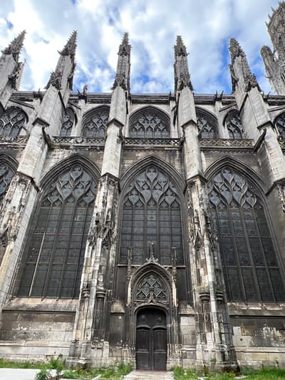 The image displays a highly detailed, upward-angled view of a section of the Flamboyant Gothic Église Saint-Maclou (Saint-Maclou Church) in Rouen, France, under a partially cloudy blue sky.

The scene focuses on the elaborate stone architecture of the church's exterior. Multiple tall, ornate stone pinnacles, characteristic of Gothic design, extend skyward from the upper structure. Below these, a series of smaller arched windows with intricate stone tracery are visible. The main body of the church features several very tall, arched windows, each dominated by complex, flame-like (flamboyant) stone tracery in their upper sections. The lower panels of these windows are glazed with dark, somewhat opaque glass, suggesting stained glass or age.

Vertical stone buttresses separate these large windows, extending from the ground level upwards. Some buttresses feature decorative elements and what appear to be gargoyles or water spouts positioned below the upper window level. The lower wall of the church is constructed from large, grey stone blocks, exhibiting signs of age, weathering, and discoloration, with some areas showing repair or lichen growth.

Centrally located at the base of the structure is a prominent, deeply recessed arched doorway. This entrance features dark, double wooden doors below an ornate stone tympanum with tracery mirroring the windows. The immediate ground in front of the church consists of a narrow strip of unkempt earth with sparse green vegetation and weeds, bordered by a lighter-colored paved surface to the left foreground. No individuals are present in the scene.