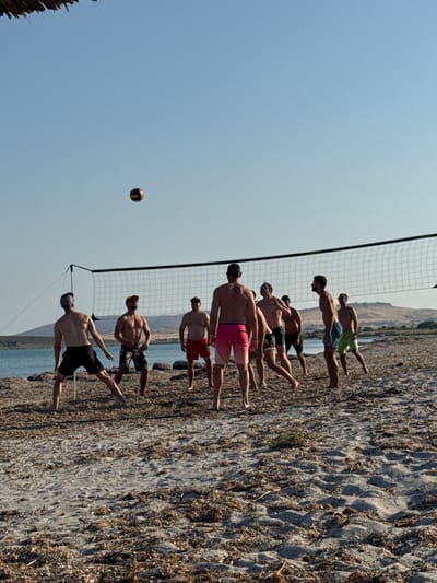 A group of men are playing beach volleyball on a rocky shore. There are at least eight men in the image, most of whom are shirtless and wearing swim trunks. The volleyball net is set up on the beach, and a volleyball is in the air, indicating an active game. The men are positioned on both sides of the net, with some in the process of hitting or receiving the ball. The ground is covered in pebbles and dry vegetation, typical of a Mediterranean coastline. In the background, the sea is visible, and beyond that, a hilly landscape. The sky is clear and blue, suggesting a sunny day, likely in the late morning or early afternoon. The overall mood appears to be one of friendly competition and recreation. The scene is set in Moudros Municipal Unit, Greece. There is no visible text in the image.
