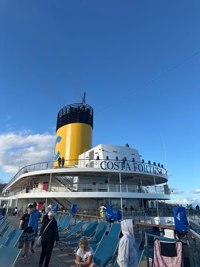 The image depicts the stern and upper decks of the cruise ship *Costa Fortuna*, captured from an elevated position on one of its lower decks. The vessel is located in the city of San Sebastián de la Gomera, Spain.
The ship's large, cylindrical funnel dominates the upper-mid section of the frame, featuring a bright yellow body with a prominent blue stylized 'C' logo on its side and a black upper section from which a light plume of exhaust is visible. Below the funnel, the ship's name, "COSTA FORTUNA," is displayed in large blue lettering on the white superstructure. Several decks with white and metallic railings are visible, populated by a number of passengers. On a deck level with the funnel, two individuals stand near the railing, observing the surroundings.
In the foreground, numerous teal-colored sun loungers are arranged on a wooden-planked deck. Several loungers are covered with blue protective sheets. Multiple individuals are present on this deck: A male subject in a black t-shirt and patterned shorts carries a blue towel while walking towards the left. A female subject in dark clothing walks behind him. Another female subject with light-colored hair, wearing a white t-shirt, sits on a sun lounger, looking downwards. To the right, an individual in a grey hooded sweatshirt stands, holding a mobile device. A patterned beach chair is visible on the far right.
The sky above is largely clear and blue with scattered white cumulus clouds on the horizon to the left. Thin black lines, indicative of rigging or overhead wires, traverse the upper portion of the sky. A distant landmass, possibly the island's terrain, is visible on the far left horizon behind the ship.