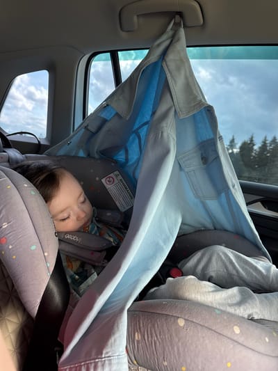 A young child sleeps in a car seat, nestled within the confines of a vehicle. A light blue denim jacket is draped over the car seat, creating a makeshift canopy. The jacket is attached to a car's interior handle, shielding the child from direct sunlight. The child is wearing a patterned shirt. The background shows a view through the car window, with a cloudy sky and trees visible. The scene appears to be a quiet moment during a car journey, possibly on a road trip. The location is identified as Staroye Martyanovo, Russia.
