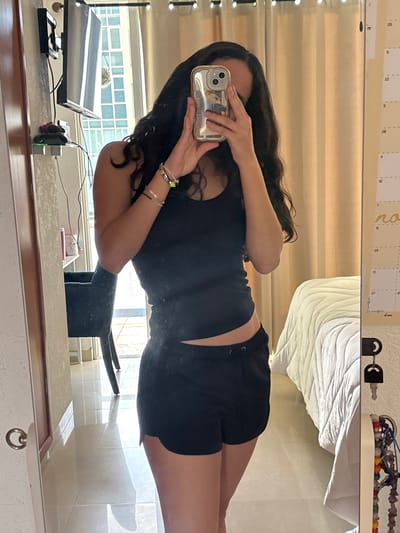 A young woman is reflected in a mirror, holding a smartphone up to take a selfie. She is wearing a black tank top and black shorts. The reflection also shows a desk with a computer monitor, keyboard, and other accessories. In the background, there is a window with a view of a building complex with large windows and a tiled roof. A calendar and other items are on a shelf above the mirror. Based on the architecture and vegetation visible through the window, the location is in the city of Boca del Río, Mexico.