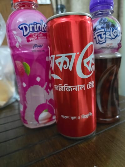 The image displays three beverage containers on a dark wooden surface. In the foreground, a red aluminum can, likely a soft drink, is prominently positioned. Its label features white text with the iconic Coca-Cola logo and Bengali script that translates to "Original Taste." To the left of the can is a pink plastic bottle containing a litchi-flavored fruit drink. This bottle also has white text indicating "Drinks," "Litchi Fruit Drinks," and "Float with Nata Jelly," along with an illustration of a litchi. To the right of the red can, a purple-capped plastic bottle holds a dark liquid, possibly a blueberry-flavored drink, with white Bengali script. The background is blurred, suggesting an indoor setting, possibly a home or a small shop in Chor Sahapur, Bangladesh. The lighting appears to be natural, casting subtle shadows on the wooden surface.