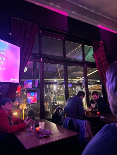 The media file captures a rotated (approximately 90 degrees clockwise) nighttime scene inside what appears to be a karaoke bar or restaurant. The foreground, positioned at the bottom right of the rotated image, shows a person wearing a red long-sleeved top and a blue baseball cap, seated at a wooden table. They are looking towards a large illuminated screen, partially visible on the left side of the rotated image. Several empty drinking glasses and a candleholder with a small flame are present on the table.
The central part of the image is dominated by a large, dark-framed window with multiple panes, separating the dimly lit interior from an outdoor area. Dark red curtains are draped along the window frame. Through the window, an outdoor patio or seating area is visible, illuminated by numerous string lights, suggesting a festive or social gathering space. Two men are seen through the window, seated at a table outdoors; one is closer to the glass, wearing a dark top, while the other in a blue shirt is further back.
On the left side of the rotated image, a large screen displays karaoke lyrics. The text "Karafuni" is visible, along with partial lyrics such as "...cos", "back", "act", and "...r". The indoor space features ambient lighting, with purple and pink hues casting a glow on the adjacent wall, adding to the vibrant atmosphere. At the very top of the rotated image, a person's hair and the top of their head are partially visible, indicating another individual is present, out of frame. The overall scene suggests a lively evening activity in a social setting.