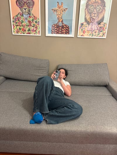 An indoor scene in Nesso, Italy, depicts a young adult female reclining on her left side on a grey fabric sectional sofa. She is dressed in a white short-sleeved t-shirt, dark blue wide-leg denim jeans, and a single bright blue slipper with white detailing on her visible right foot. Both hands grasp a grey smartphone, equipped with a white circular magnetic accessory, and her gaze is directed at the screen. Above the sofa, mounted on a plain light brown wall, are three framed art prints featuring anthropomorphic animals wearing suits and eyeglasses: from left to right, a leopard in a floral suit, a giraffe in a patterned suit, and a lion in a floral suit with a pink cravat. The room is uniformly lit.