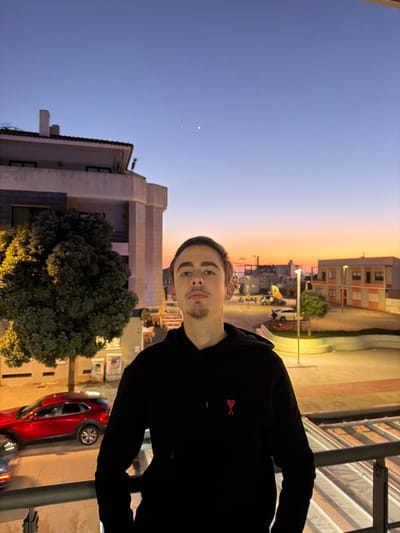 A young male stands on a balcony railing at dusk in Gafanha da Nazaré, Portugal. The individual, in his late teens or early twenties, has short dark hair, light-colored eyes, a thin mustache, and a small goatee. He wears a black hooded sweatshirt with a small, red heart-shaped logo (Ami de Cœur) on the left chest. He is positioned in the lower center of the frame, looking directly forward.
Behind the individual, an urban street scene unfolds during twilight. To the left, a multi-story building with a light-colored facade and a reddish-brown tiled roof is partially visible, with a large, illuminated green tree in front of it. A red SUV is parked or moving on the street below the tree, with another vehicle partially visible. To the right, additional residential or commercial buildings are illuminated by streetlights. The horizon features a band of orange and pink light from the sunset, transitioning to a deep blue sky overhead. Two distinct, faint points of light, consistent with celestial bodies, are visible in the upper right quadrant of the sky. The foreground is partially obscured by a dark metal balcony railing. Artificial lights from buildings, streetlights, and vehicles are visible throughout the scene.