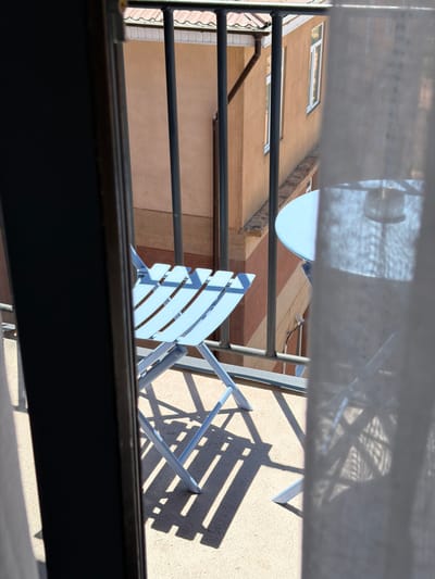 The image is a close-up, low-angle shot of a balcony in Kaji-Say, Kyrgyzstan. In the foreground, a light blue metal folding chair is visible on the balcony floor, casting long shadows. To the right, a portion of a round, light blue metal table is seen, also casting shadows. A sheer white curtain partially obscures the right side of the image, and a dark vertical element, possibly a doorframe, is on the left. In the background, a building with terracotta-colored walls and white-framed windows is visible beyond a metal railing. The scene appears to be set on a sunny day.