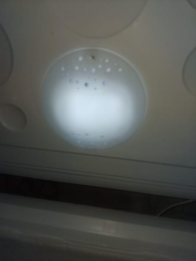 The image shows a close-up, low-angle view of a ceiling light fixture. The fixture appears to be circular with a translucent cover that diffuses the light. Small, circular patterns or indentations are visible on the cover, some of which are darker and appear to be openings or recesses. A small, dark circular object, possibly a screw or a decorative element, is visible in the center of the fixture. The surrounding ceiling is a plain, light-colored surface. Below the ceiling, horizontal lines suggest the edge of a structure or furniture, and a dark recessed area is visible, possibly indicating a shadowed space or the top of a cabinet. The overall scene is indoor and dimly lit, with the primary light source being the ceiling fixture itself. The location is the city of Haveli Jhangranwali, Pakistan.