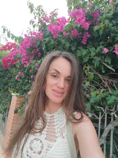The image is a selfie of a woman with long, brown hair and braces. She is smiling and looking at the camera. She is wearing a cream-colored crocheted top. Behind her is a wall of vibrant pink and red bougainvillea flowers, with green leaves. The background also includes a section of a brick pillar, a metal fence, and some dry branches. The scene appears to be outdoors, possibly in a residential area in El Campello, Spain, given the presence of bougainvillea, common in Mediterranean regions.