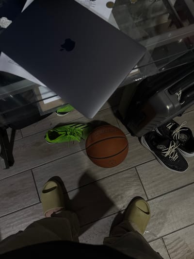 The image captures a casual scene on a tiled floor. A grey laptop sits on a glass table, accompanied by papers underneath. Nearby, a suitcase stands upright, next to a pair of black and white sneakers. An orange basketball and a neon green running shoe are also present on the floor. A person wearing beige slides is partly visible on the right side of the frame. The photograph was likely taken indoors. Without additional information, identifying the exact location in Chiapa de Corzo, Mexico is not possible.