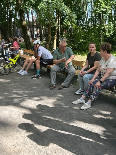 A group of five adults and one child are resting on a wooden bench in a park setting in Dubrovitsy, Russia. The bench is situated on a paved area with dappled sunlight filtering through trees.
On the left, a child sits on a yellow bicycle. Beside the child, a woman in a pink t-shirt sits on the bench.
Next to her, a man wearing a white cycling jersey, black shorts with "PAS" written on them, and a white helmet is seated. He is looking down, with his hand on his chin.
To his right, a man in a green polo shirt and grey trousers is also seated. He appears to be talking, with his hands gesturing.
Next to him, a woman in a dark t-shirt and jeans is seated, looking towards the right.
On the far right, a woman in a light-colored floral shirt and patterned trousers is seated. She is looking to her left.
The environment is lush with mature trees providing shade. Shadows of the trees and possibly the people are cast on the ground.