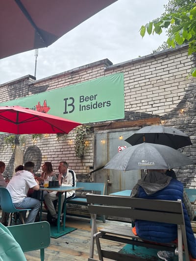 The image depicts an outdoor seating area of a establishment, possibly a cafe or restaurant, in Komarovo, Russia. The scene is characterized by a slightly overcast sky, suggesting inclement weather, which is further evidenced by the presence of two open umbrellas. One umbrella is black and appears to be positioned above a group of people seated at a table. The second umbrella, also black, has text in Russian that appears to say "РИВЬЕРА" (Riviera) and is shielding individuals on a bench.
Several people are visible, seated at tables and on benches. In the foreground, a group of four individuals are seated at a turquoise table. One person, wearing a white t-shirt and blue jeans, is facing away from the camera. Next to them, another individual is also partially visible. Across the table, a woman with blonde hair is looking towards a man in a white and black jacket who is also seated at the table. There is a glass of amber liquid, likely beer, on the table. Further back, more people are obscured by umbrellas and seating.
The environment includes a brick wall adorned with a large green banner featuring a logo and text in Russian and English, reading "Beer Insiders". There are hanging plants and string lights affixed to the wall. The seating area is constructed with wooden planks for the floor and various types of seating, including turquoise chairs and benches. A red umbrella is also visible, angled over the seating area from the left side of the frame. The overall impression is a functional outdoor space designed to accommodate patrons, even in less than ideal weather conditions.