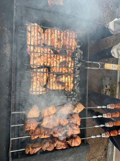 The scene depicts an outdoor grilling operation in Заозерное Снт, Russia. A rectangular, dark metal brazier holds glowing and ash-covered charcoal, from which thick white smoke emanates. In the upper portion of the brazier, a metal grilling basket contains numerous pieces of light meat, consistent with poultry, exhibiting browning and some charring. The wooden handle of this basket is held by a partially visible individual on the right, whose lower legs are clad in dark trousers and light footwear, with a grey fabric sleeve also present. Below the grilling basket, four metal skewers, each with a twisted shaft and a decorative wooden handle, are laid across the brazier. These skewers hold multiple large, cubed pieces of red meat, similarly browned from cooking. The surrounding ground is unpaved, consisting of dry earth.