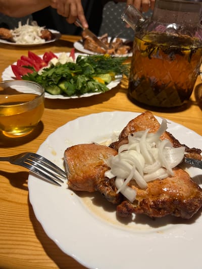 This close-up, high-angle shot captures a dinner scene on a wooden table. The main focus is a white plate filled with grilled meat, likely kebabs, topped with thinly sliced white onions. A fork rests on the plate, suggesting a meal in progress. To the left, a glass pitcher, filled with a golden liquid and ice, is being poured, with a person's hand visible holding it. Next to the pitcher is a plate of fresh vegetables, including chopped tomatoes, cucumber slices, and fresh herbs like dill and parsley. Another glass of the same golden liquid sits near the vegetable plate. In the lower left, another plate with food, possibly more kebabs and shredded cabbage, is partially visible, along with a hand holding a knife. The lighting suggests it is likely nighttime, possibly indoors, and the overall atmosphere appears casual and convivial. There is no visible text in the image.