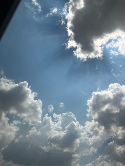 This is a photograph of the sky, taken from a low angle, looking upwards. The main subjects are clouds and the sky itself. The clouds are varied in appearance, with some being dark and dense, suggesting they may be rain clouds, while others are bright and wispy, illuminated by sunlight. The sky is a clear blue, with rays of sunlight beaming through gaps in the clouds, creating a dramatic effect. The lighting suggests it is daytime, likely late morning or early afternoon, given the intensity of the sun. There are no people or man-made objects clearly visible, and no text is present in the image. The overall scene evokes a sense of grandeur and the power of nature, with a mix of dramatic and serene elements. The perspective and the framing of the clouds create a sense of depth and vastness.