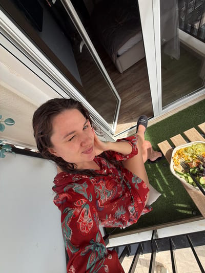 A woman with wet hair is on a balcony in Glifada, Greece. She is wearing a red patterned dress and black slippers. She is seated on a white cushion, with a salad on a table covered in artificial turf. The balcony has a black railing. A partially open glass door reveals a bed indoors.