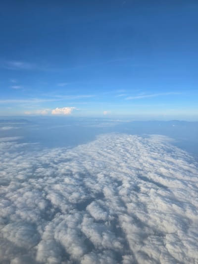 The image shows a high-altitude view of clouds from an airplane. The sky is a clear, bright blue, with some wispy white clouds scattered in the distance. Below, a vast expanse of puffy white clouds stretches as far as the eye can see, creating a textured, undulating surface. In the far distance, faint outlines of mountains or landmasses can be seen through the haze. The perspective suggests the viewer is looking down at the cloud layer, with the horizon line a distant feature. There are no people, objects, or actions visible in this aerial landscape. The environment is entirely natural, consisting of the atmosphere and cloud formations. The image does not provide any specific information that would identify its location within Tanohanoe, Indonesia.