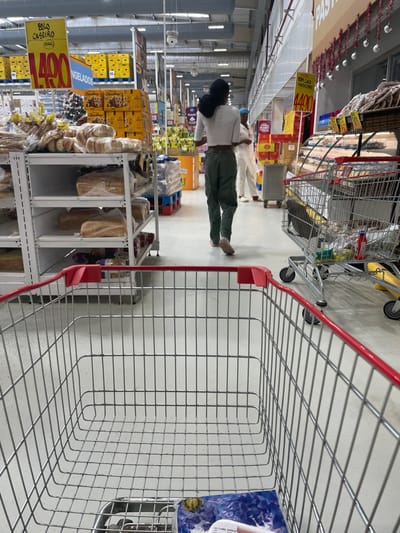 An image captured from the perspective of a shopping cart shows a supermarket aisle. In the foreground, the empty wire basket and red handle of a shopping cart are prominent. A woman with dark hair, wearing a light-colored top and dark green pants, walks away from the camera down the wide aisle. She is in motion, with her right foot slightly lifted. Further down the aisle, two other individuals in light clothing are partially visible.
The supermarket is brightly lit with artificial lighting. On the left side of the aisle, numerous shelves are stocked with various products. There are stacks of boxed goods, possibly cookies or biscuits, and plastic-wrapped bread loaves on lower shelves. A large yellow sign on the left reads "BOLO CASEIRO 1.490" (Homemade Cake 1.490). Below it, another sign partially visible states "CONGELADOS" (Frozen). Further down the aisle, another yellow sign advertises "TOM GALO 1.400" (Gallo Tomato 1.400). The prices, featuring values like 1.490 and 1.400, suggest a local currency. On the right side of the aisle, beyond the foreground cart, more shelves and products can be seen. Another shopping cart, appearing partially loaded, is situated behind the woman. The activity depicted is grocery shopping in a well-stocked and orderly supermarket environment in Luanda, Angola.