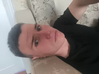 The image shows a close-up, low-angle, selfie-style photograph of a young person lying on a couch. The person has short, dark hair and is wearing a black t-shirt. Their head is tilted back, and their gaze is directed towards the camera. The background consists of a patterned wallpaper and a portion of the couch. The lighting is dim, and the image is slightly out of focus. The context provided about Gremyachinsk, Russia, does not appear to be reflected in the visual content of the image itself.