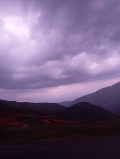 The image captures a twilight landscape with a dramatic sky over rolling hills. The sky is filled with heavy, dark purple clouds, illuminated from above by a bright, diffused light source, suggesting either the setting sun or significant cloud cover. In the middle ground, rounded, dark hills are visible, with some areas appearing to have a reddish hue, possibly due to vegetation or soil type. The foreground is dark and out of focus, possibly representing a road or plain. The scene conveys a sense of moody, atmospheric weather conditions in a natural, undeveloped environment. This scene could be found in the mountainous regions surrounding Taldyk, Kyrgyzstan, known for its dramatic skies and rugged terrain.
