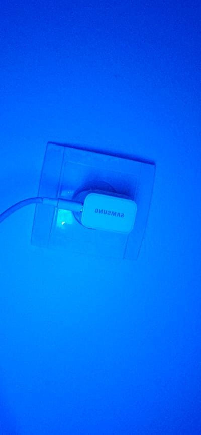 An electrical outlet is shown in the image, illuminated by a blue light. A white Samsung charging adapter is plugged into the outlet, with a white cable extending from it. The outlet and adapter are mounted on a pale surface, likely a wall. The surrounding environment is entirely saturated with a deep blue hue. There are no people or other objects visible in the scene. The location is the city of Yangikishak, Uzbekistan.