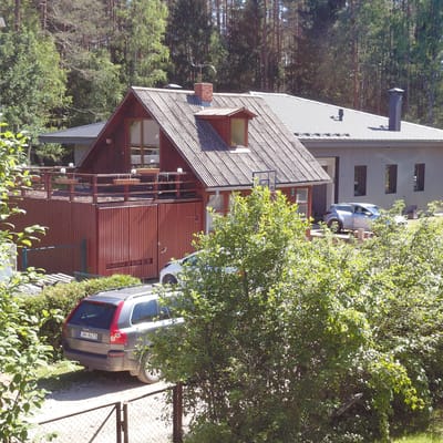 The image depicts a residential scene in Skuķīši, Latvia, under what appears to be bright daylight, possibly midday, with clear weather. The foreground is dominated by lush green foliage, which partially obscures the view of a multi-story wooden house. The house has a distinctive shingled roof and a dark red wooden exterior on its lower levels. Visible architectural features include several windows, a small dormer window, a balcony or deck area with wooden railings, and a chimney.
To the left of the house, a lighter-colored, modern-looking building is visible, with a gray, ribbed siding. A silver car is parked in front of this building. Further to the right, a darker gray SUV is parked on a gravel driveway next to a chain-link fence. The rear of the SUV clearly displays the license plate "JH-8473". The overall setting suggests a tranquil, possibly rural or suburban, environment with dense trees and greenery surrounding the properties. There are no people visible in the image, and the scene appears to be static, with no particular activity taking place other than the presence of the parked vehicles. The image is captured from a slightly elevated perspective, looking down towards the house and driveway.