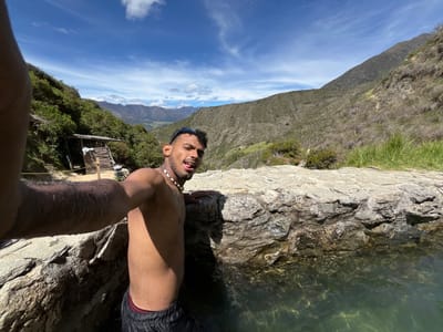 A man is taking a selfie in a natural hot spring, located in the mountains outside of Mucurubá, Venezuela. The man, who is shirtless and wearing black swim trunks, is standing in the hot spring with his arm extended, holding the camera. The hot spring is surrounded by large rocks and lush green vegetation. In the background, the mountains of the Venezuelan Andes rise majestically under a bright blue sky with scattered white clouds. A small wooden structure, possibly a changing room or shelter, can be seen on the hillside to the left. The water in the hot spring is clear and inviting, with sunlight reflecting off its surface. The overall impression is one of relaxation and connection with nature.