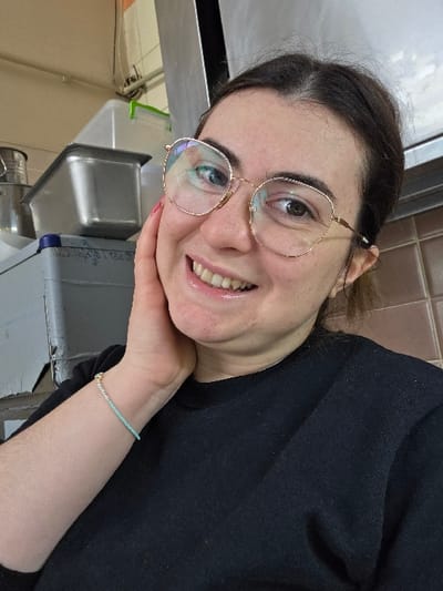 An indoor, close-up image captures a female individual with dark hair and gold-rimmed glasses smiling directly at the camera, her left hand gently touching her cheek. She wears a black long-sleeved shirt and a light blue beaded bracelet on her left wrist. The background, visible primarily in the upper left and right, consists of an industrial kitchen or food preparation area. Visible objects include stainless steel containers, white plastic storage bins (one with a green lid), and a grey plastic crate with a blue rim. Walls appear to be light-colored, with some visible wiring, and a vertical metallic surface is present on the right. This scene is located in the city of Gevgelija, North Macedonia.