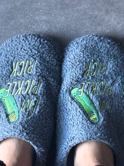 A top-down photographic view displays a pair of feet wearing matching grey-blue, fuzzy slippers on a dark grey, textured surface, likely a floor, within a location in Limeil-Brévannes, France. Each slipper is embroidered with lime-green text reading "I'M PICKLE RICK" and features a prominent embroidered patch depicting a cartoon green pickle character with facial features. The bare forefeet of an individual, exhibiting a fair skin tone, are visible protruding from the front openings of the slippers. No further subjects, objects, or actions are discernable.
