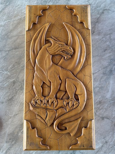 A rectangular, golden-brown wooden carving depicts a dragon. The dragon faces left, its mouth open, revealing sharp teeth. It has wings and clawed feet. The background around the dragon is textured with small, polygonal shapes. The edges of the rectangular wooden frame feature stylized, wavy cutouts. The carving is placed on a grey, marble-patterned surface. There is no indication of the location being Таскино, Russia, or any people in the scene.