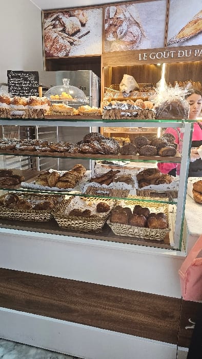 This image depicts the interior of a bakery located in Bir Mourad Rais, Algeria. A multi-tiered glass display case dominates the foreground and midground, showcasing a variety of baked goods.
The display case's top shelf presents an assortment of pastries, cakes, and a large, round item wrapped in clear plastic, with some yellow pastries covered by a glass cloche. The middle shelf holds numerous darker-colored pastries, some dusted with powdered sugar, arranged on white paper liners. The bottom shelf contains several woven wicker baskets filled with bread rolls and other small baked items.
In the background, a wooden sign above the display reads "LE GOÛT DU PAIN." Three framed photographs of baked goods are mounted on the wall. To the left, a blackboard sign, partially obscured, displays prices. A stainless steel commercial appliance, bearing the name "isa," is visible behind the counter.
A young woman with dark hair, wearing a pink top and carrying a light pink bag on her arm, is partially visible on the right side of the image, positioned in front of the display case. Her head is turned downwards or away from the camera. The counter beneath the display case is white with a dark wood panel, and the floor is tiled.