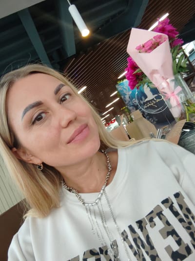 A medium-angle, eye-level selfie is captured from the shoulders up. A blonde woman with light skin and brown eyes is looking towards the camera, slightly to the left. She has dark eyebrows and is wearing makeup, including eyeliner and lipstick. She is wearing a white sweatshirt with a leopard print design across the chest, and multiple silver necklaces. A black clover-shaped earring with a gold border is visible on her left ear. In the background, a bouquet of pink roses and fuchsia peonies is displayed in a clear vase. The environment appears to be indoors, with a modern ceiling structure featuring wooden panels and exposed beams. A cylindrical light fixture hangs from the ceiling. The overall impression is a snapshot taken in an interior setting, possibly a cafe or a shop. The specific location in the city of Amursk, Russia, is not discernible from the visual information provided.