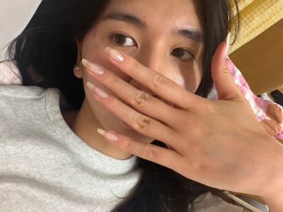 A person with long, dark hair is lying down, partially obscuring their face with their hand. The person's eyes are visible, with dark irises. The person is wearing a light gray, ribbed top. The fingers of the hand are elongated with clear nails. The setting suggests an indoor location. There are elements of a pink and white patterned object to the left, and a light-colored object towards the lower right corner. The location in the city of Kawasaki, Japan cannot be determined from this image.