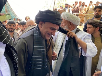 The image shows a group of men in a crowd in Zaker-E Sharif, Afghanistan, at the coordinates 31.547 latitude and 65.761 longitude. The main subject is a man wearing a black turban and a long black jacket with white stripes, who is being presented with a black shawl by a man in a white robe. The man in the white robe is holding the shawl with his left hand and gently placing it around the neck of the man in the black turban. Other men stand behind and around them, some looking on and others partially obscured by the main subjects. The background shows a crowd of people, some of whom are standing on a metal railing. The scene appears to be taking place in a public outdoor space. The lighting is bright and the scene is well-lit. There is a smartphone in the foreground partially visible, suggesting someone may be taking a photo. The men are dressed in traditional Afghan clothing and the image likely shows a religious or cultural event.