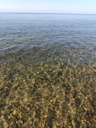 The image depicts a clear, shallow body of water with a pebble-strewn bottom, likely a shoreline. The water is calm, with gentle ripples distorting the view of the submerged rocks. Sunlight penetrates the water, creating a shimmering effect and highlighting the varied colors and textures of the pebbles. The horizon line is visible in the upper portion of the image, separating the water from a pale, cloudless sky. There are no people or man-made objects visible. The scene is located in Kurskoe, Kyrgyzstan, a region known for its natural landscapes.