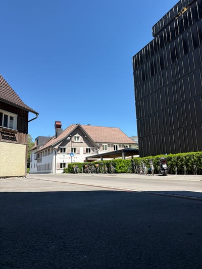 The image captures a street scene in the city of Hard, Austria, on a clear, sunny day. In the foreground, a dark asphalt road takes up the lower portion of the frame, with a distinct shadow cast from an unseen object on the left. To the left of the road, a building with a light yellow textured facade and a brown tiled roof is partially visible. Adjacent to this, a white building with brown shutters and a red-tiled roof is situated. A sign on this building reads "Gasthaus KATH'R". In front of the Gasthaus KATH'R, a line of bicycles and a scooter are parked along a low, dense green hedge. To the right of the Gasthaus KATH'R, a modern, tall black building with vertical paneling dominates the right side of the image, reaching towards the bright blue sky. The overall environment suggests a commercial or residential area with a mix of traditional and contemporary architecture.