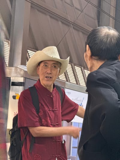 In Tiaodeng, China, two men are standing near a metro map. The man on the left wears a straw cowboy hat and a maroon collared shirt with frog buttons. He has a black backpack. The man on the right wears a black jacket and has thinning hair. They appear to be engaged in conversation or discussion, with the man in the cowboy hat gesturing towards the map. The environment suggests an indoor public transportation hub, likely a metro station, with a brightly lit ceiling and tiled walls.